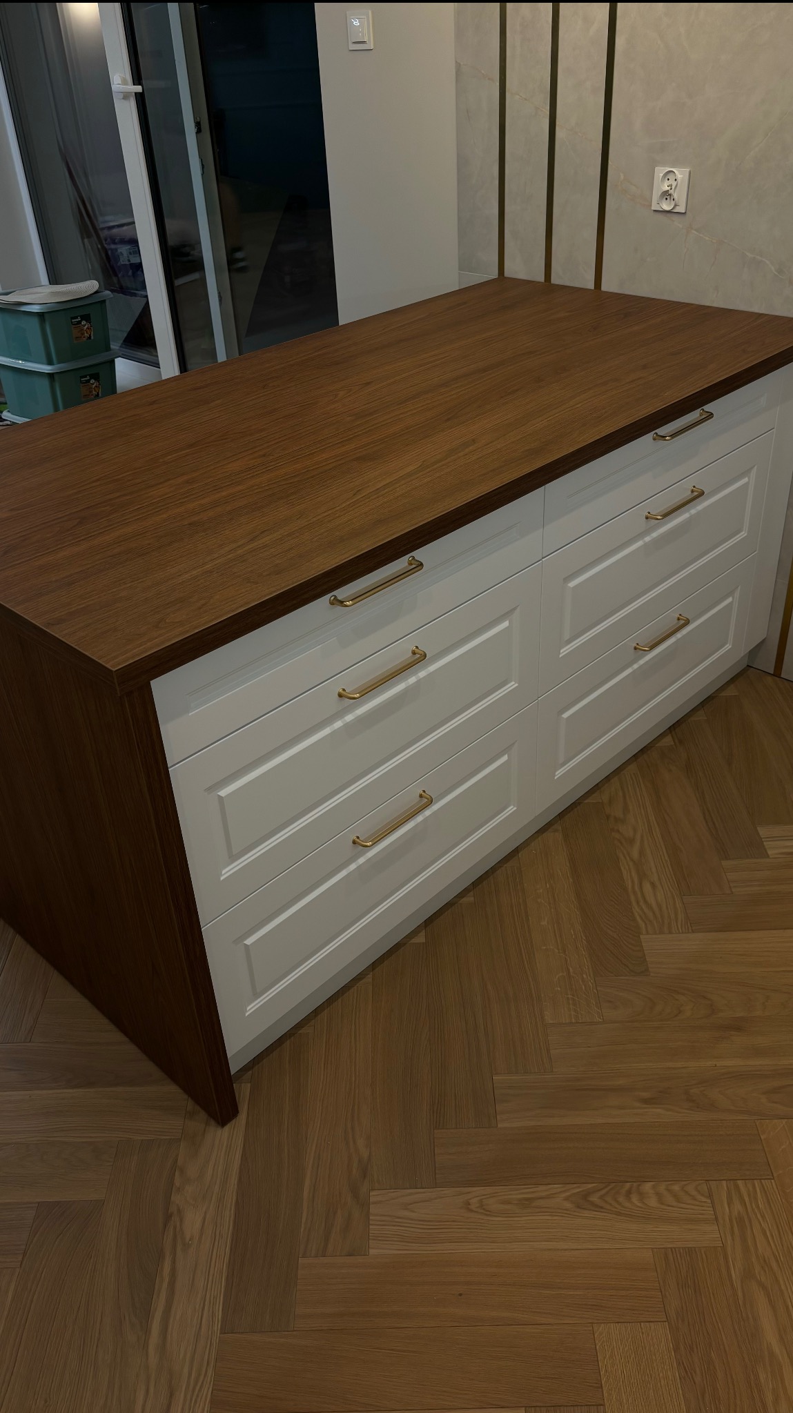 Wysuwana komoda z brązowym blatem i białymi frontami szuflad z złotymi uchwytami, na podłodze z parkietem ułożonym w jodełkę.