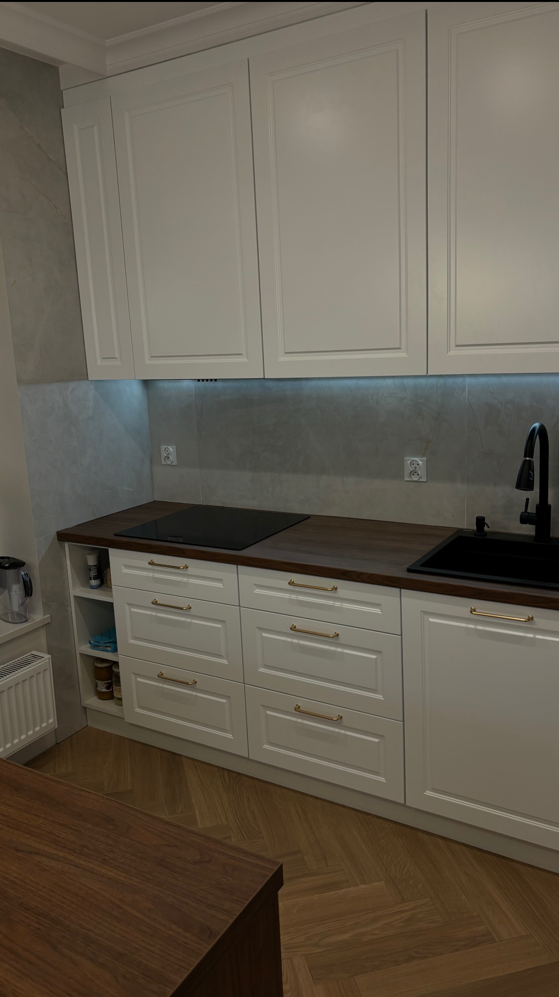 Jasna kuchnia na wymiar z drewnianym blatem i złotymi uchwytami, płyta indukcyjna i czarny zlew. Podświetlenie LED pod szafkami górnymi. Parkiet w jodełkę na podłodze.