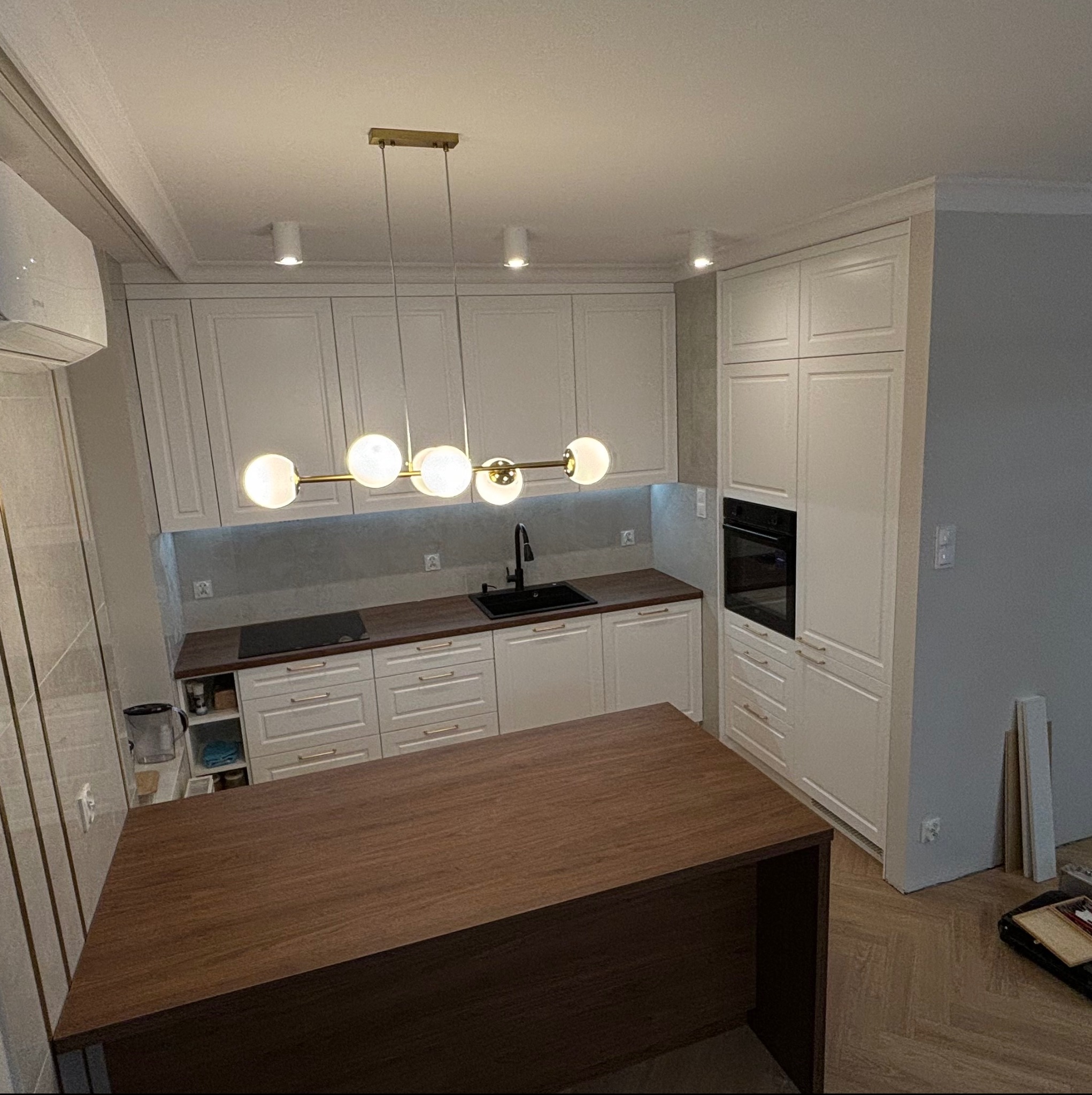 Nowoczesna kuchnia z białymi frontami, drewnianym blatem i wyspą. Nad blatem wisząca lampa z kulistymi kloszami. Na podłodze parkiet ułożony w jodełkę.