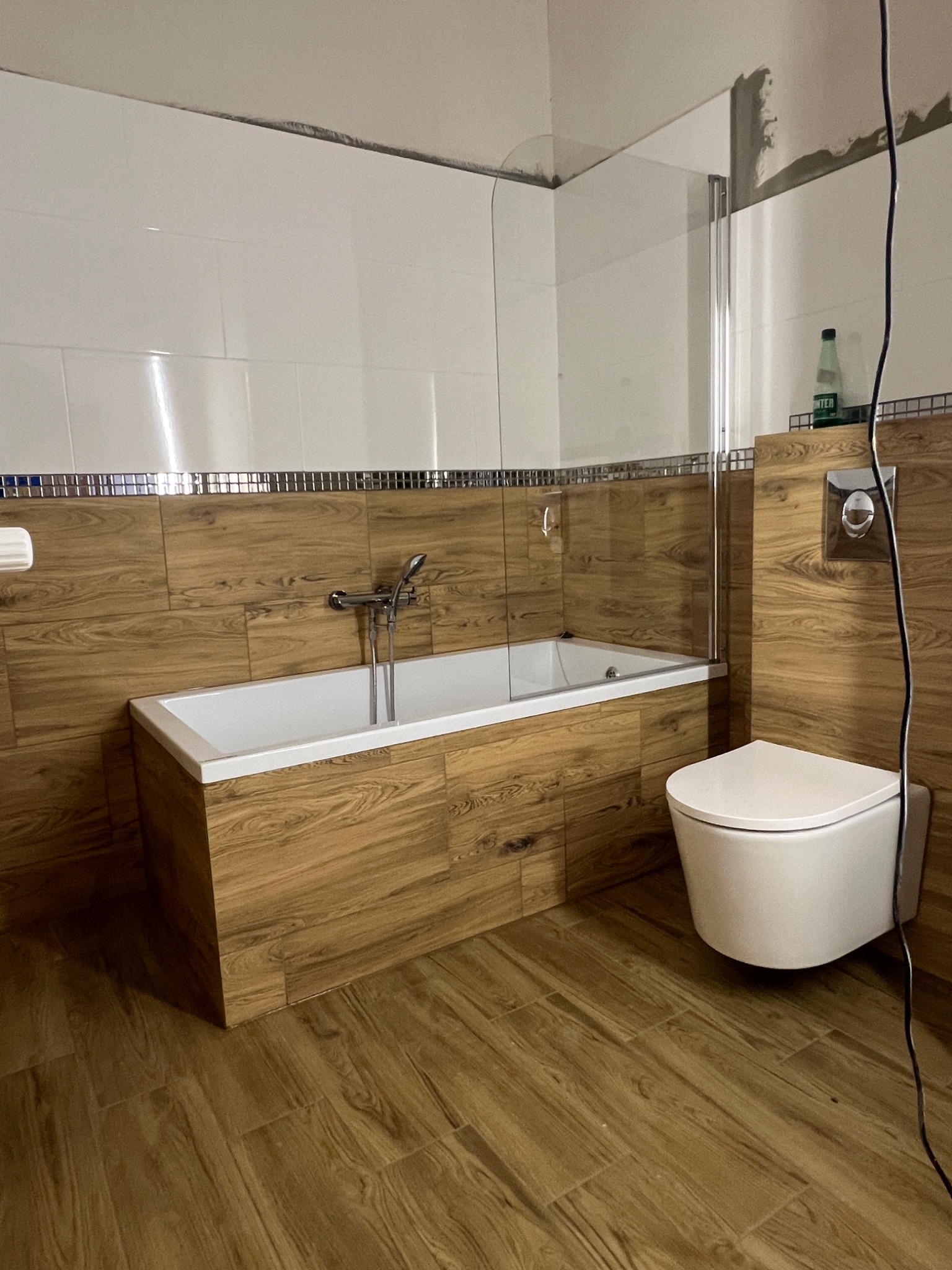 Wykończona łazienka z wanną obudowaną płytkami imitującymi drewno, toaletą podwieszaną i szklanym parawanem nawannowym. Widoczne fugi i listwa dekoracyjna.