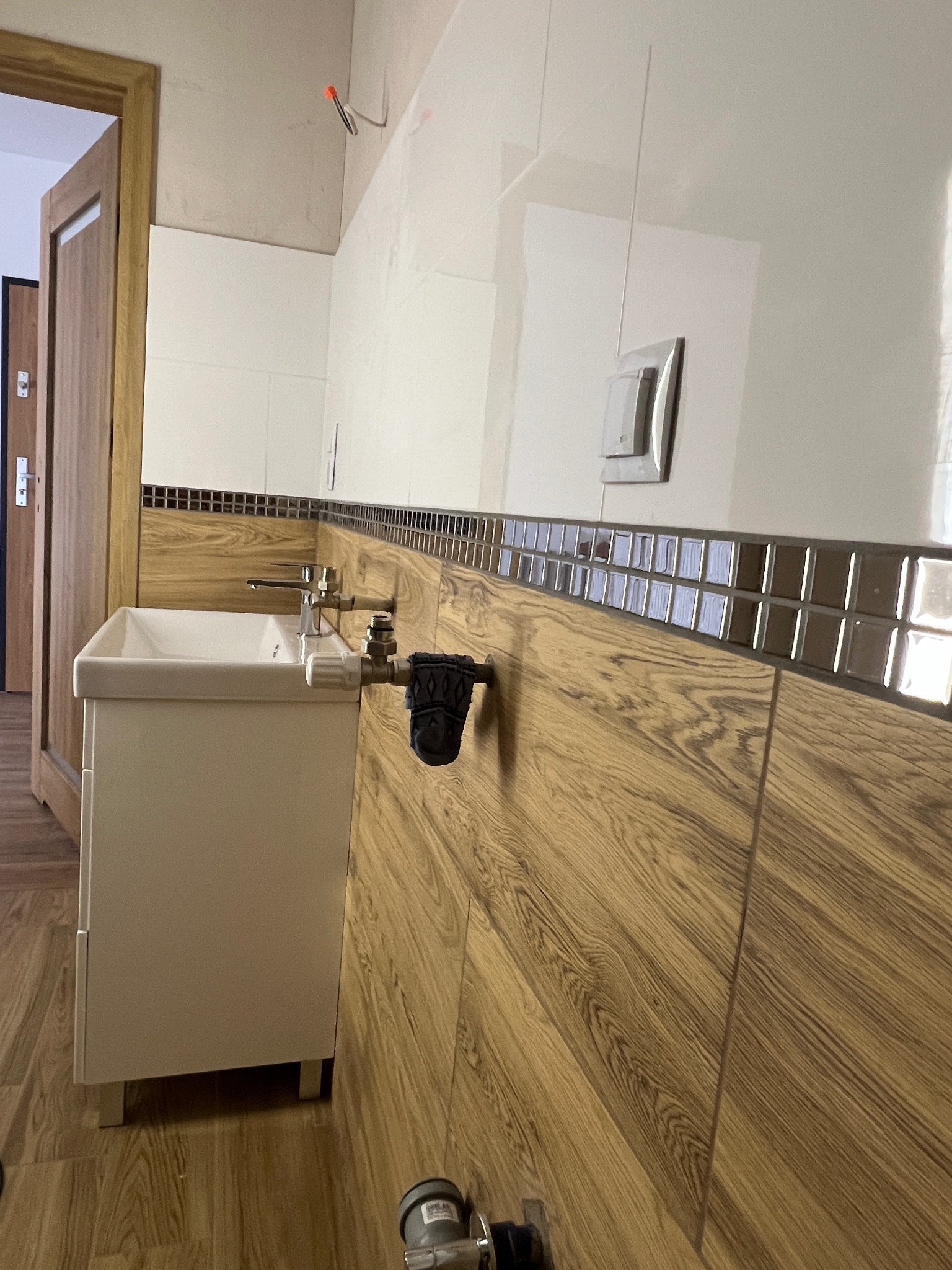 Wykończona łazienka z umywalką i armaturą, drewnopodobne płytki na ścianach i podłodze, mozaika dekoracyjna. Widoczny fragment drzwi i instalacji elektrycznej.