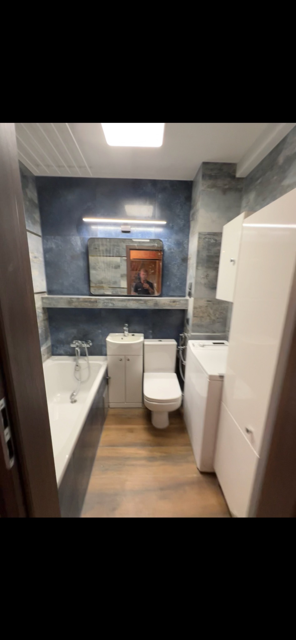 Wykończona łazienka z wanną, toaletą, umywalką i pralką. Na ścianach płytki w odcieniach szarości i niebieskiego. Lustro z oświetleniem nad umywalką. Podłoga imitująca drewno.