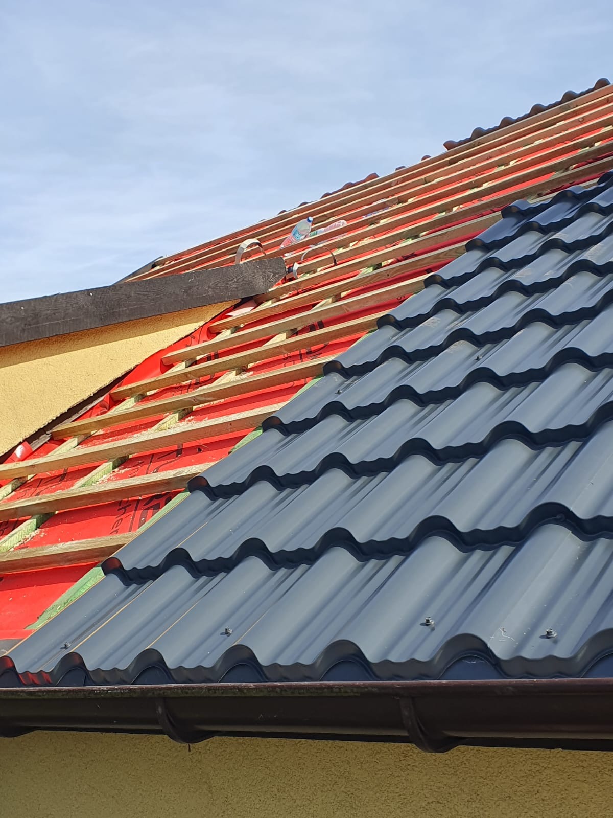 Częściowo pokryty dach z widoczną konstrukcją więźby, membraną i nową, ciemną blachodachówką w falisty wzór, oraz zamontowaną rynną.