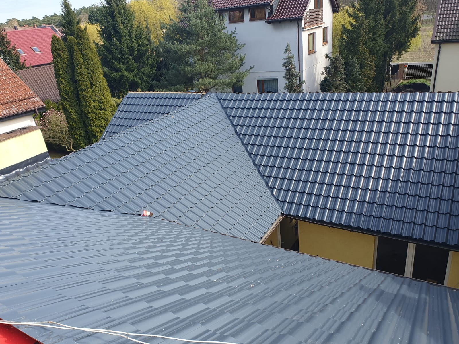 Nowy, grafitowy dach z metalowej dachówki modułowej, widok z góry na tle zabudowy jednorodzinnej.