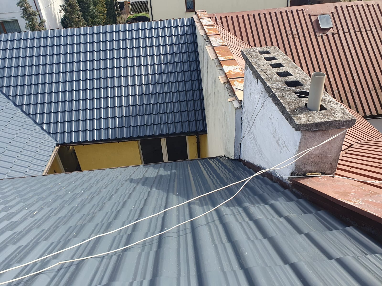 Widok z góry na fragment dachu z blachy w odcieniach szarości, sąsiadujący z dachem z czerwonej dachówki i kominem z wystającą rurą. Widoczne przewody elektryczne biegnące wzdłuż połączenia dachu...