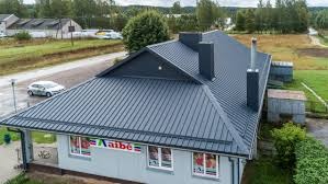 Budynek z nowym, ciemnoszarym dachem z blachy trapezowej, widok z góry, z kominami z tego samego materiału, otoczony zielenią i drogą.