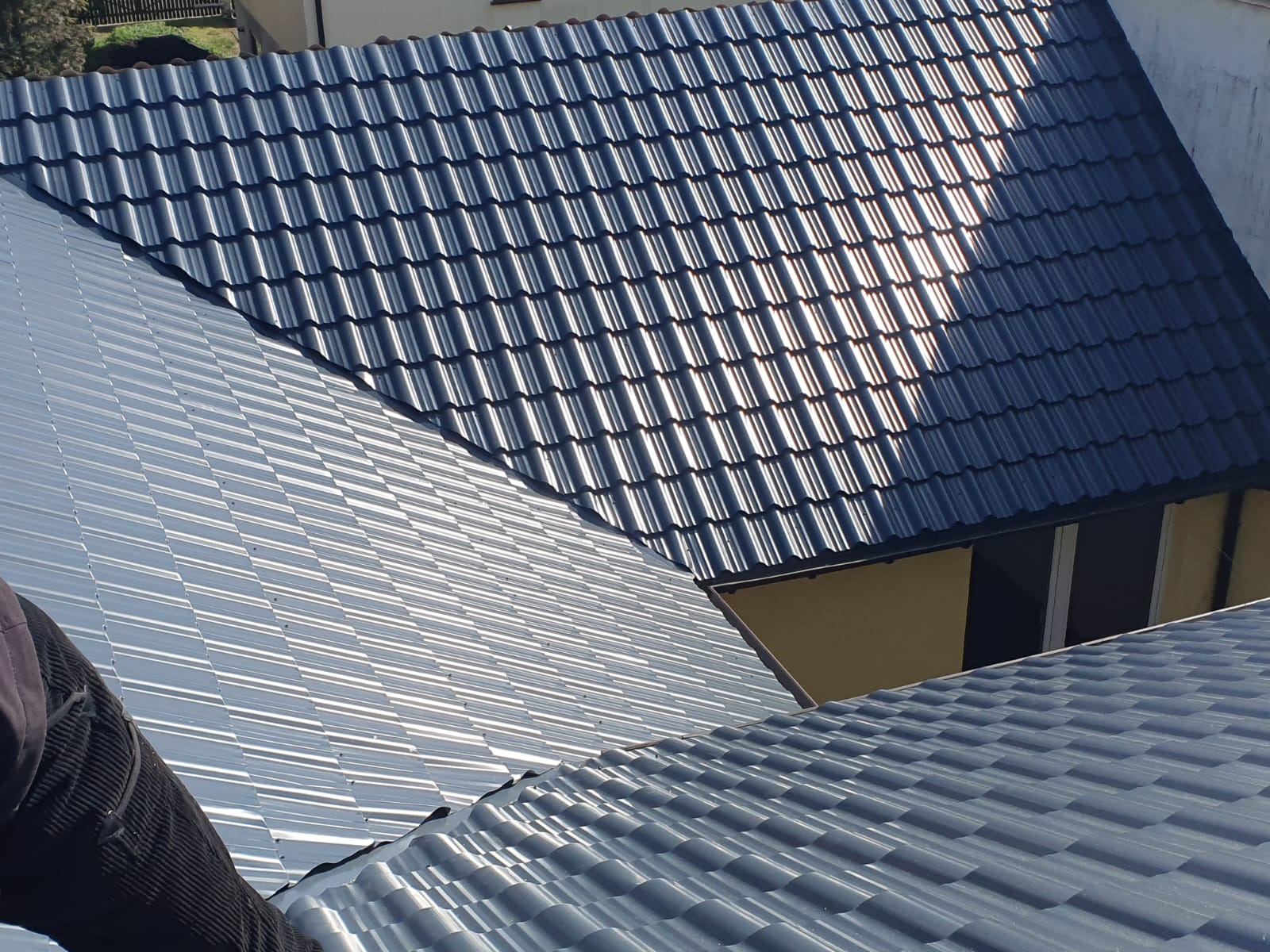 Fragment dachu z widocznym połączeniem blachy trapezowej i dachówki ceramicznej w kolorze grafitowym, z refleksami słonecznymi.