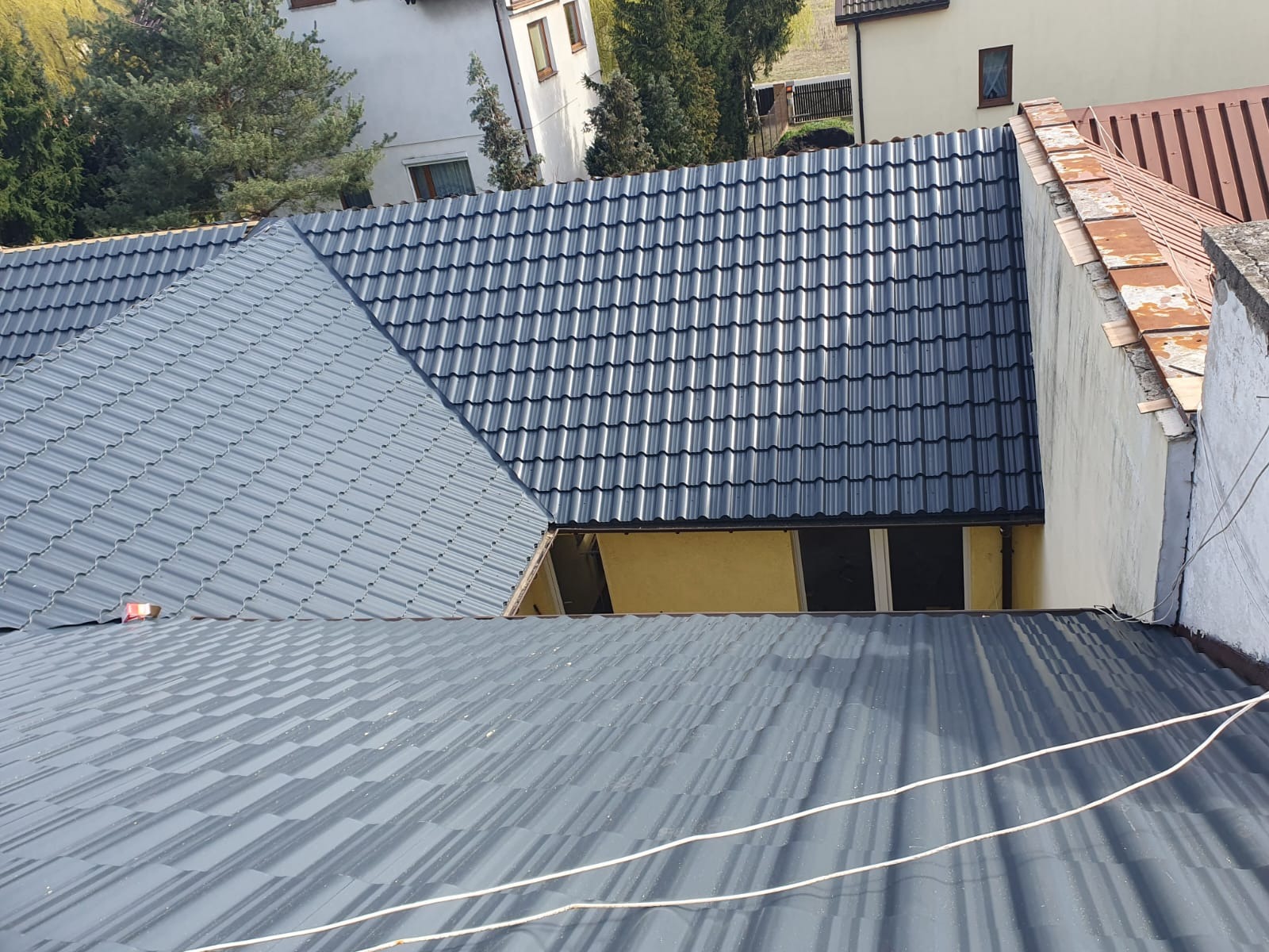 Widok z góry na fragment dachu z szarej blachy trapezowej i ciemnoszarej dachówki modułowej, widoczne okno dachowe w ścianie szczytowej i fragment sąsiedniego budynku z pomarańczową dachówką.