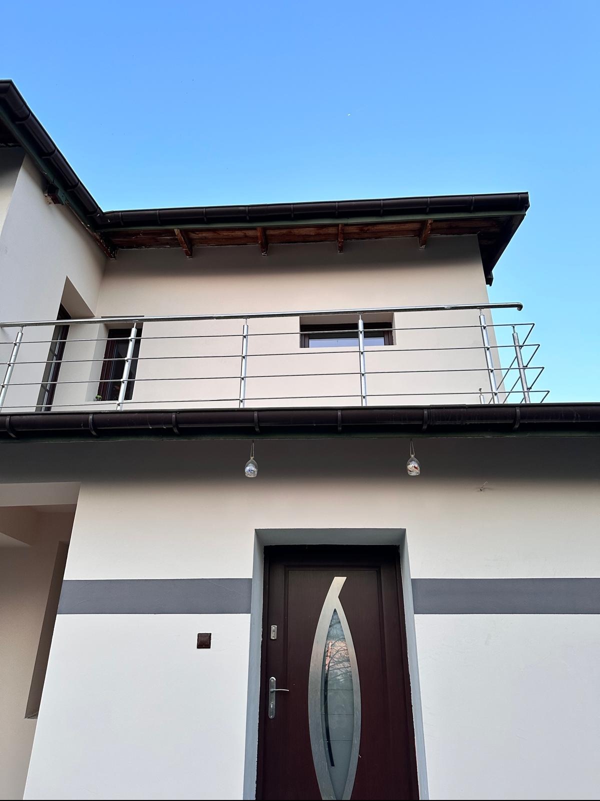 Elewacja domu po remoncie: białe ściany z szarym pasem, ciemne drzwi z szybą w kształcie łzy, balkon z metalową balustradą, widok od dołu na tle błękitnego nieba.