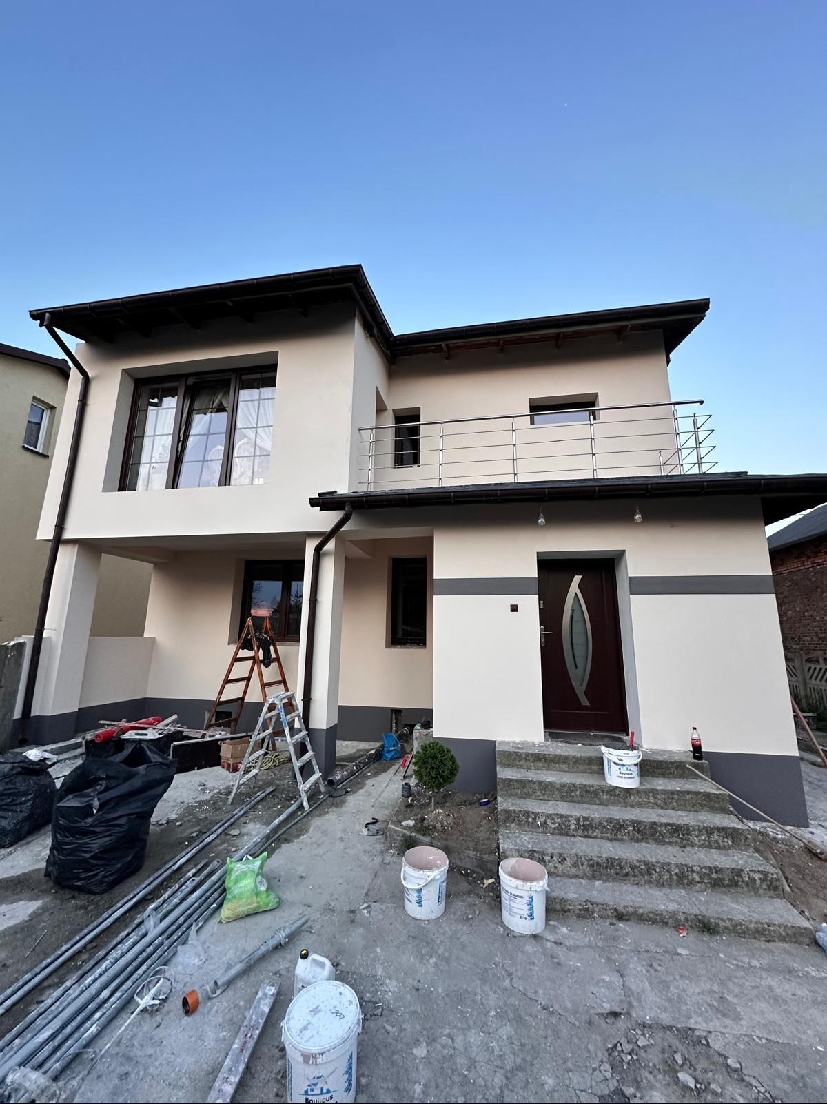 Dwupiętrowy dom po remoncie elewacji, z widocznymi detalami wykończenia, takimi jak balustrada balkonu, schody wejściowe i elementy dekoracyjne. Widoczne drabiny i farby.