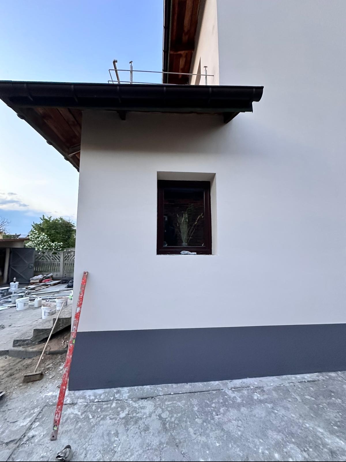 Ściana budynku po malowaniu z widoczną dolną, ciemniejszą partią. Przy ścianie stoi poziomica. W tle widoczne puszki po farbie i narzędzia.