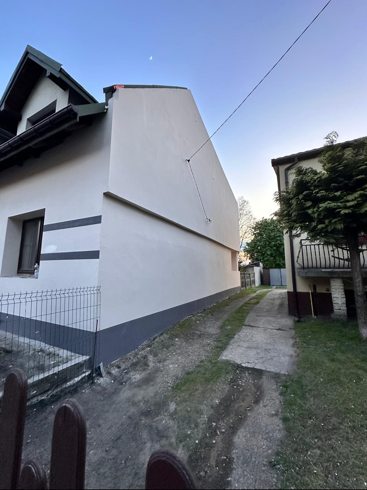 Boczny widok domu po ociepleniu elewacji, z widoczną świeżą, białą fasadą i szarym cokołem. W tle fragment posesji i sąsiedni budynek.