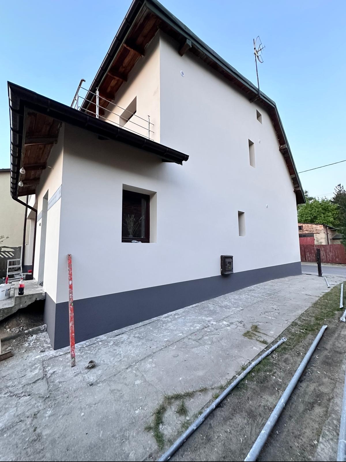 Świeżo pomalowana elewacja domu jednorodzinnego w jasnych barwach z ciemnoszarym cokołem, widok z boku, elementy rusztowania leżą na ziemi.