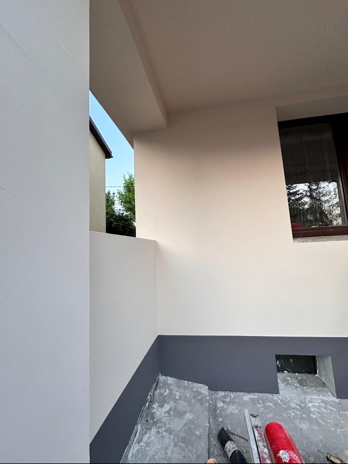 Ocieplona elewacja budynku z pomalowaną na szaro dolną częścią. Widoczne narzędzia malarskie. Okno z firanką odbija drzewa. Prace w toku.