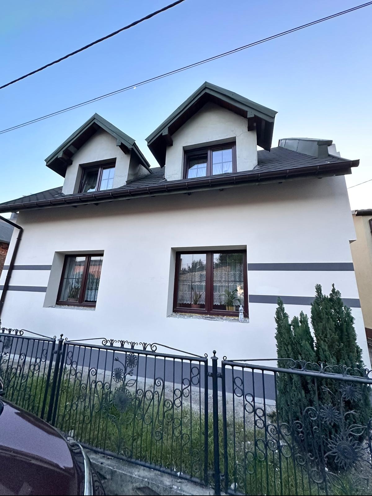 Dom jednorodzinny po remoncie elewacji, z dwoma oknami dachowymi i dekoracyjnym pasem na ścianie. Widoczne ogrodzenie i zieleń, błękitne niebo w tle.