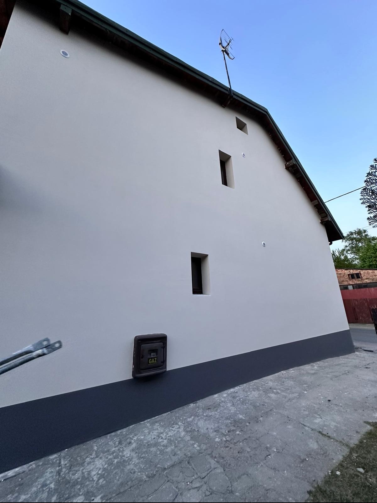 Świeżo pomalowana elewacja budynku z widoczną skrzynką gazową, dwoma małymi oknami i anteną na dachu, w słoneczny dzień.