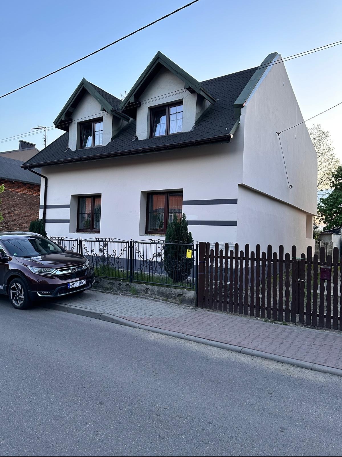 Dom jednorodzinny z jasną elewacją, ciemnym dachem i dwoma oknami dachowymi. Widoczny fragment ulicy i samochód. Ujęcie z poziomu ulicy, w tle inne budynki.