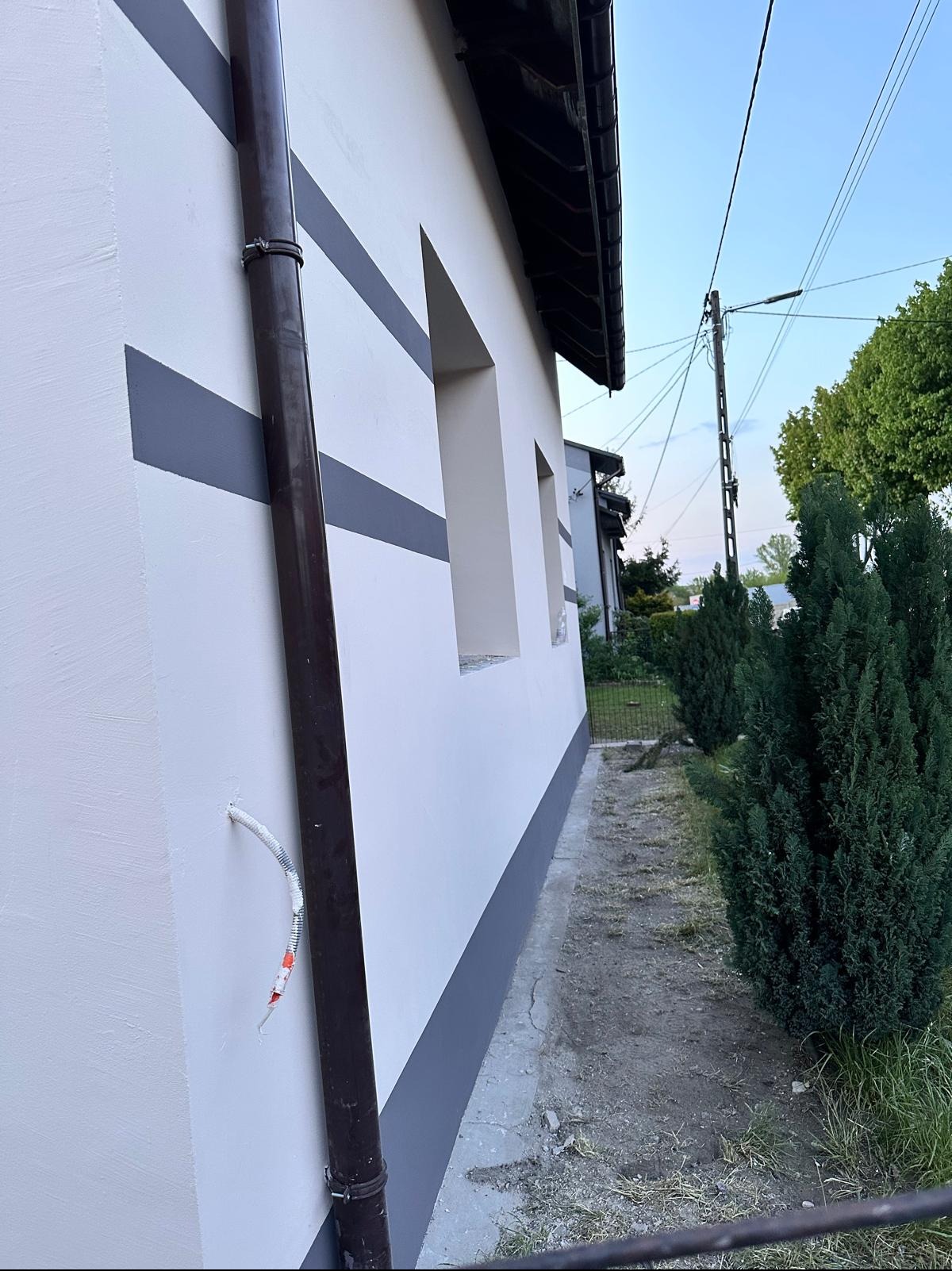 Ocieplona ściana budynku z brązową rynną i poziomymi pasami. Widoczny fragment elewacji, okna i fragment ogrodu. Na ścianie wystający kabel w peszlu.
