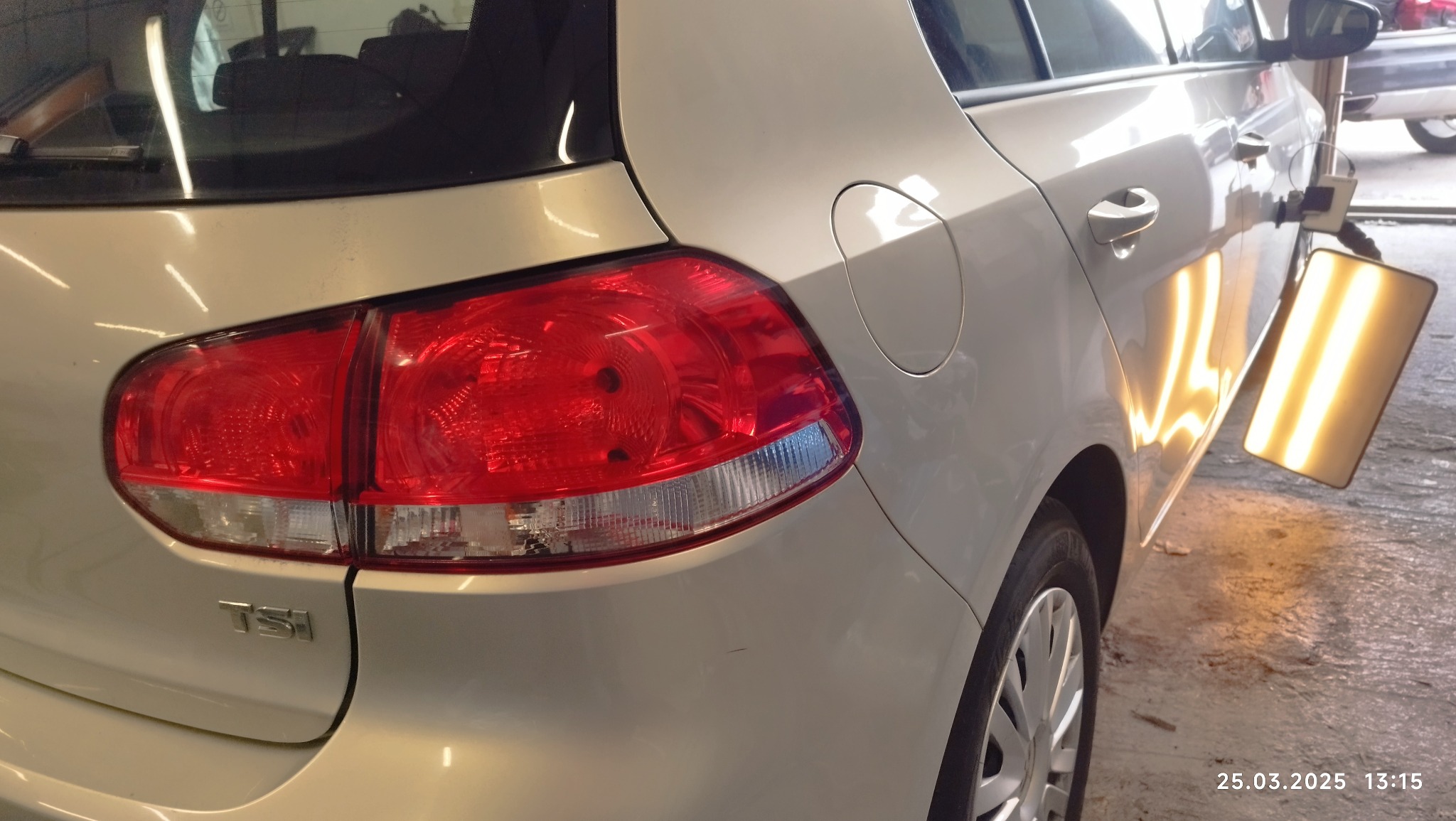 Biały samochód marki Volkswagen Golf z widocznym tylnym światłem i narzędziem do wyciągania wgnieceń na karoserii, odbijającym się w lakierze, na tle posadzki warsztatowej.