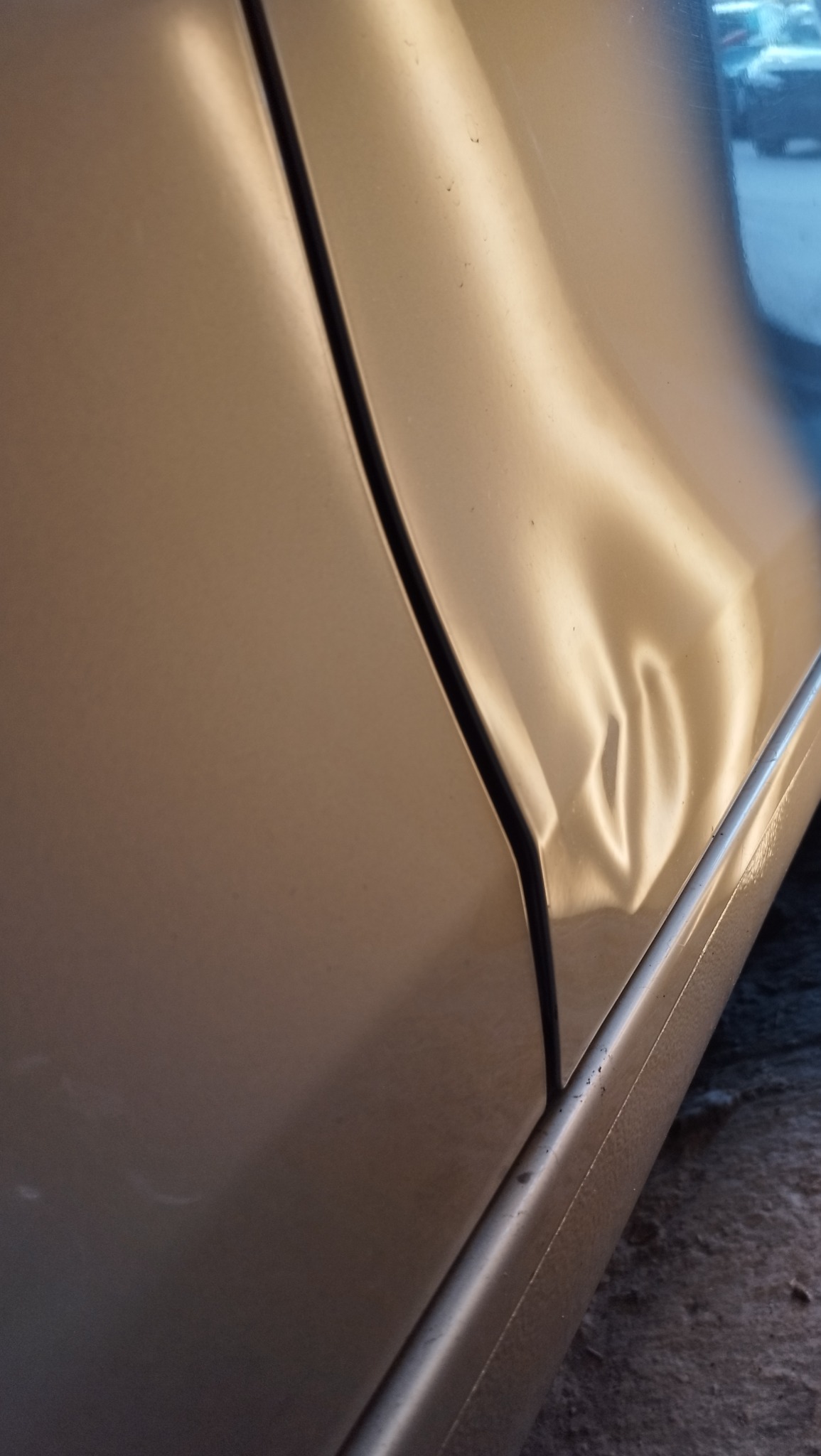 Wgniecenie na drzwiach samochodu w kolorze szampańskim, widoczne z bliska, zarysowane światłem.