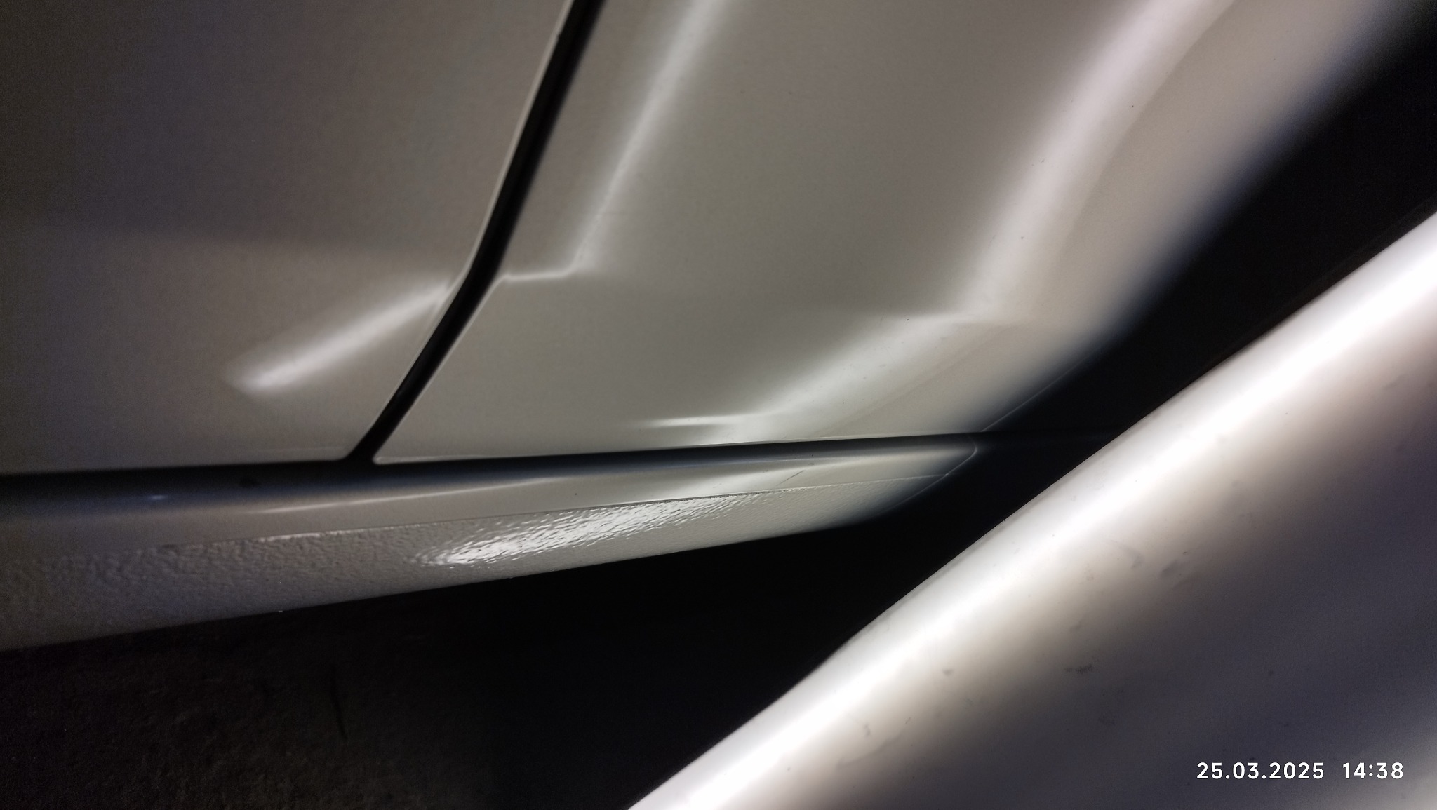 Fragment karoserii białego samochodu z widocznym wgnieceniem i odbiciem narzędzia blacharskiego, sugerujące naprawę uszkodzenia.