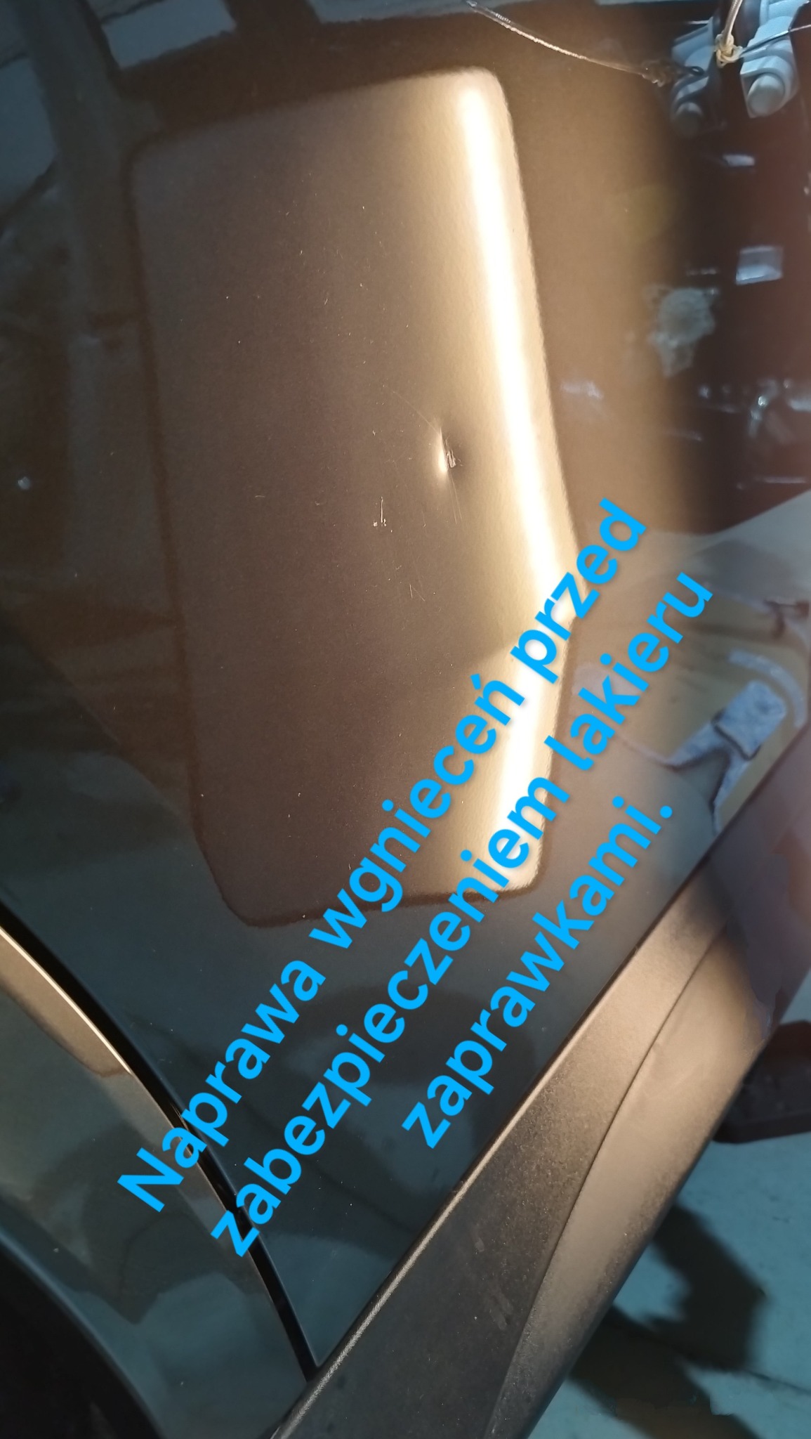 Błyszcząca, brązowa karoseria samochodu z widocznym wgnieceniem przed naprawą, zabezpieczona niebieskimi zaprawkami.