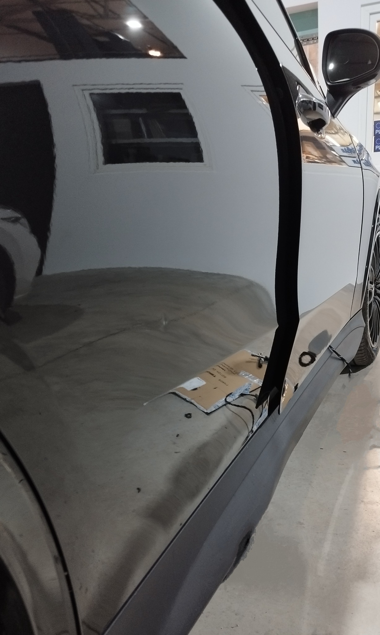 Błyszcząca, czarna karoseria samochodu z otwartymi drzwiami, widoczne narzędzia i materiały przygotowane do naprawy progu, odbicie okna w lakierze.