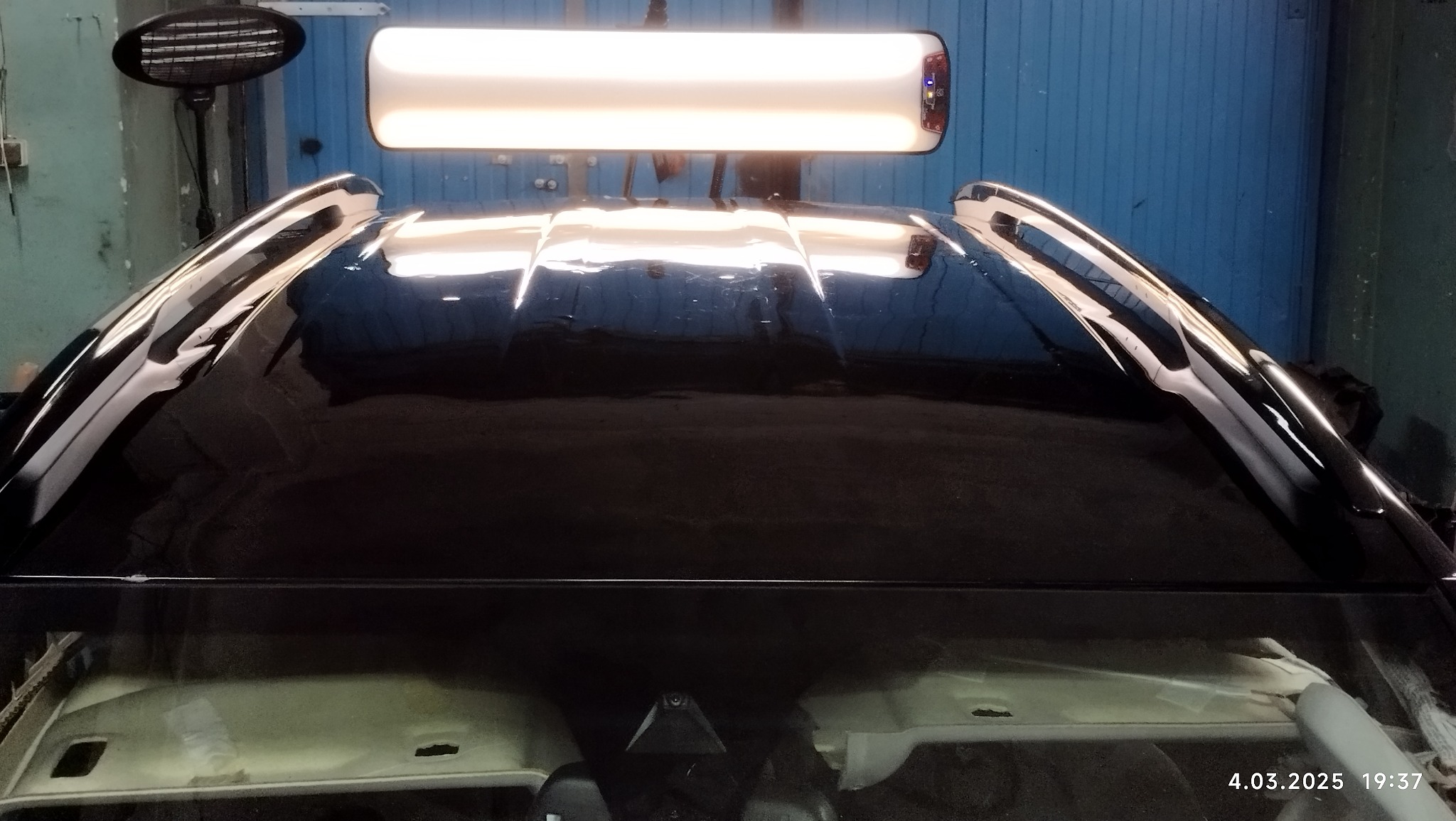 Czarny samochód pod lampą inspekcyjną w warsztacie, widoczne odbicia światła na lakierze karoserii, detale wnętrza przez przednią szybę.