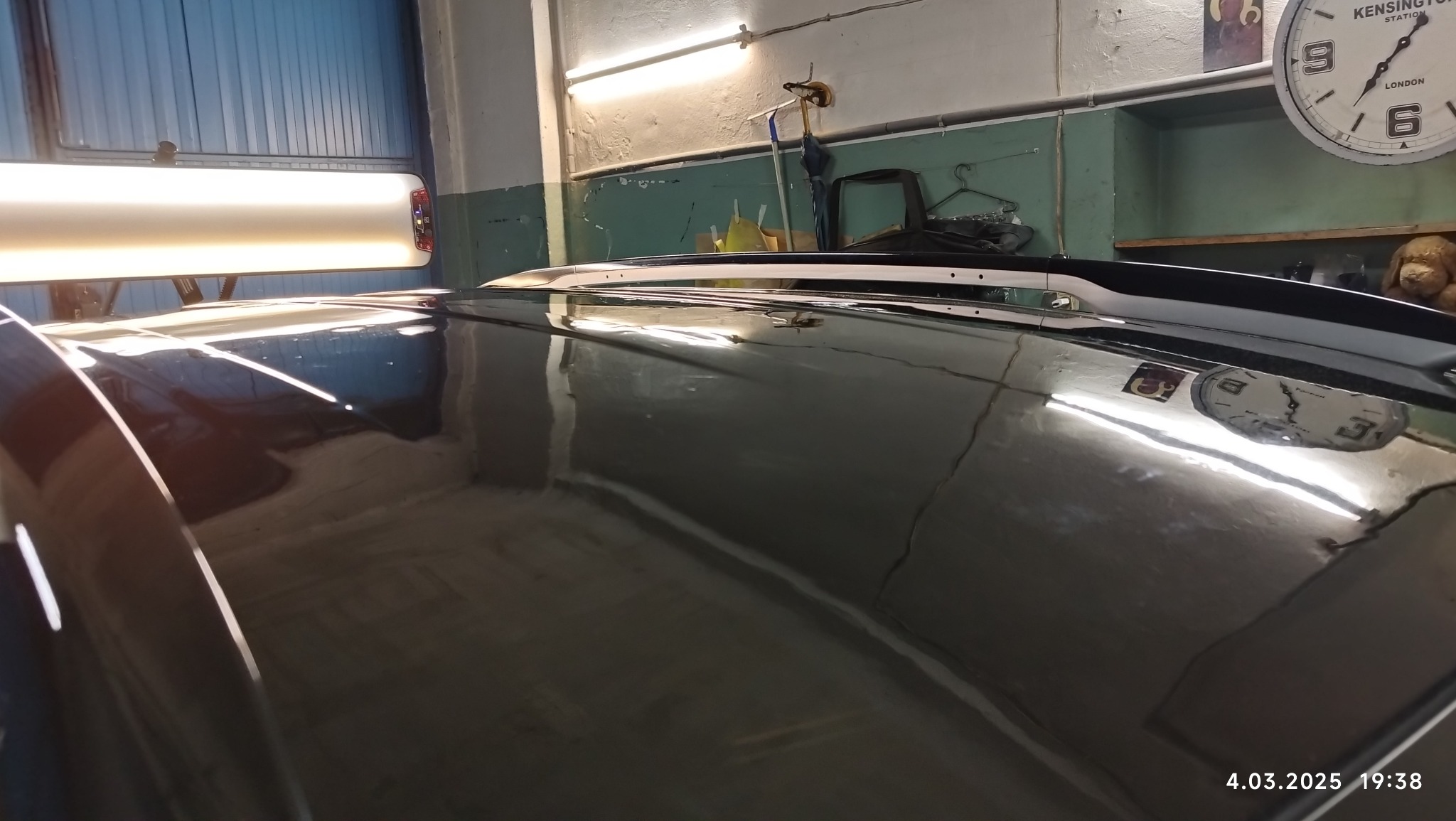 Błyszcząca, czarna karoseria samochodu pod lampą warsztatową, widoczne odbicia zegara i innych przedmiotów w lakierze. Dach samochodu z relingami. Na ścianie widoczny zegar.