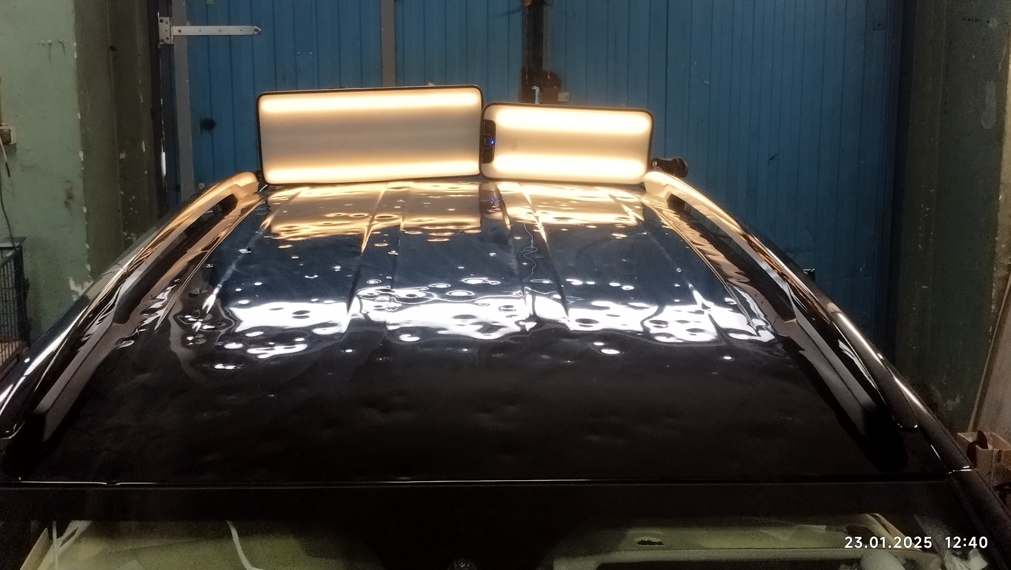 Czarne auto z widocznymi wgnieceniami na dachu, oświetlone dwoma lampami inspekcyjnymi podczas usuwania wgnieceń metodą PDR.