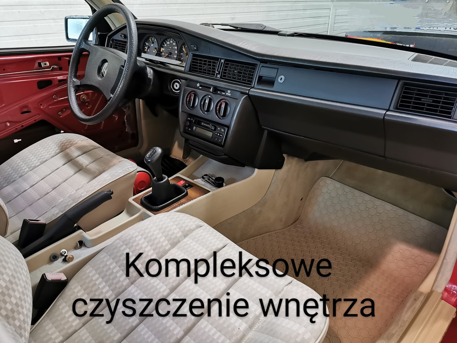 Wnętrze klasycznego samochodu Mercedes-Benz W201 z widocznymi elementami deski rozdzielczej, kierownicy, foteli z jasną tapicerką w kratę oraz fragmentami czerwonej karoserii podczas prac...