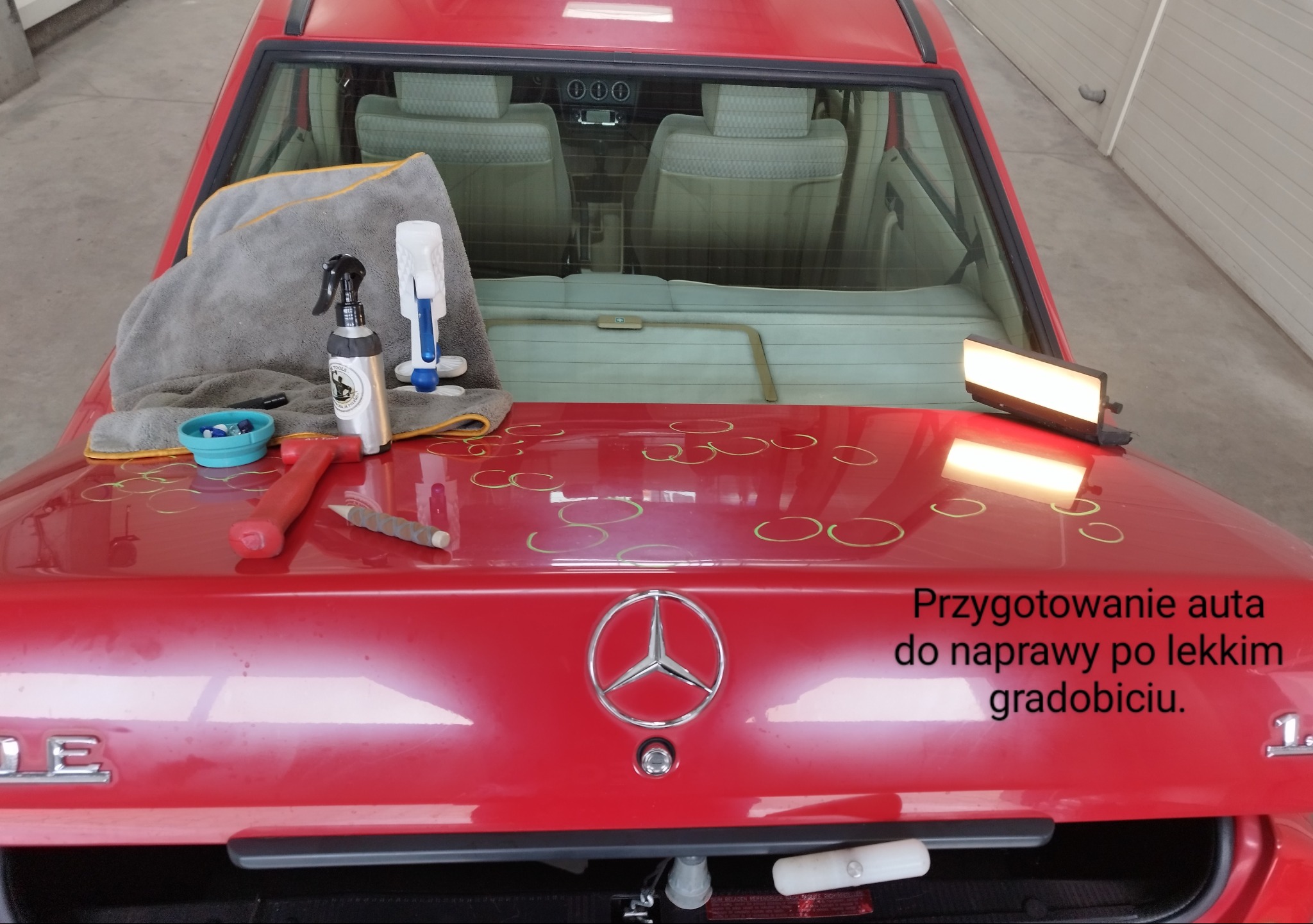 Czerwony Mercedes przygotowany do naprawy po gradobiciu, z zaznaczonymi miejscami uszkodzeń, narzędziami i lampą inspekcyjną na klapie bagażnika.