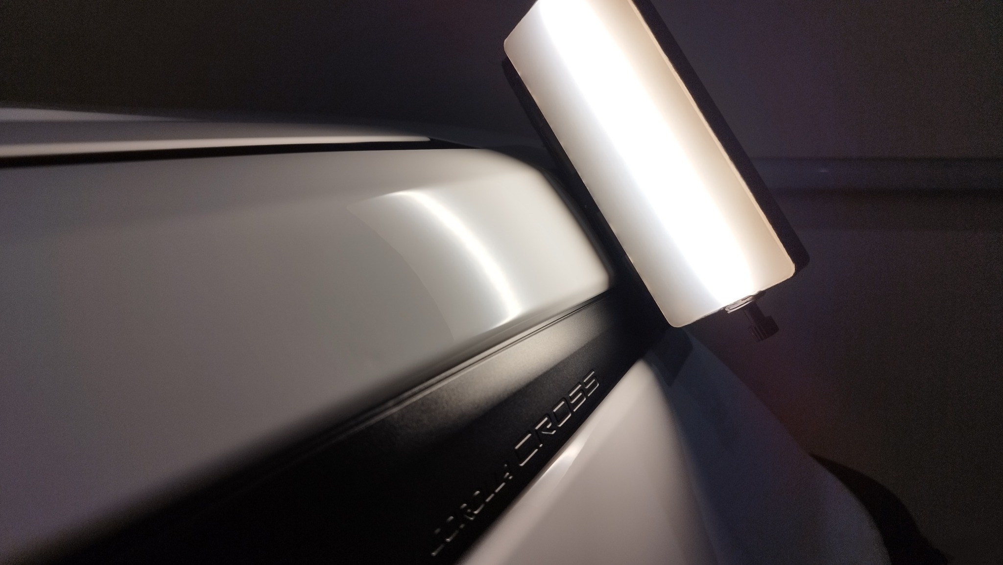 Błyszczący biały fragment karoserii samochodu z czarnym elementem z napisem 'COROLLA CROSS', oświetlony prostokątną lampą inspekcyjną.