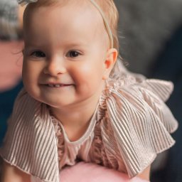 Patrycja Niedzielska - Uśmiechnięta dziewczynka w sukience w paski, z opaską z kwiatkiem, oparta na pluszowej śwince. Portret w ciepłych barwach, miękkie światło.