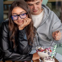 Patrycja Niedzielska - Uśmiechnięta kobieta w okularach i skórzanej kurtce siedzi z mężczyzną przy stoliku z deserami: ciastem, lodami i kawą, w jasnym oświetleniu kawiarni.