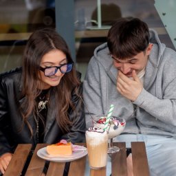 Patrycja Niedzielska - Uśmiechnięta para siedzi przy drewnianym stoliku w kawiarni, zajadając deser i pijąc kawę. Dziewczyna w okularach i chłopak w bluzie z kapturem.