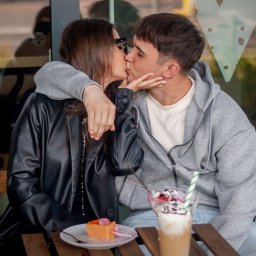 Patrycja Niedzielska - Zakochana para całuje się przy stoliku kawiarnianym, obok desery i napoje. Ona w czarnej kurtce, on w szarej bluzie. Ujęcie z bliska, na pierwszym planie jedzenie.