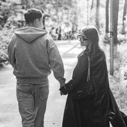 Patrycja Niedzielska - Czarno-białe zdjęcie pary trzymającej się za ręce podczas spaceru w parku, widok z tyłu, z delikatnym światłem słonecznym prześwitującym przez drzewa.