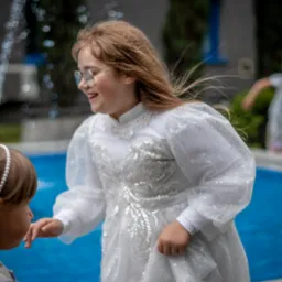 Dziewczynka w białej, błyszczącej sukience i okularach śmieje się w pobliżu fontanny. Widoczna druga dziewczynka z perłami we włosach.