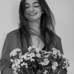 Patrycja Niedzielska - Uśmiechnięta brunetka w marynarce trzyma bukiet białych kwiatów na rozmytym tle, czarno-białe zdjęcie.