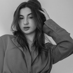 Patrycja Niedzielska - Portret kobiety w marynarce z paskiem, ujęcie od dołu, czarno-białe zdjęcie z delikatnym ziarnem, ręka we włosach, subtelny makijaż i biżuteria.