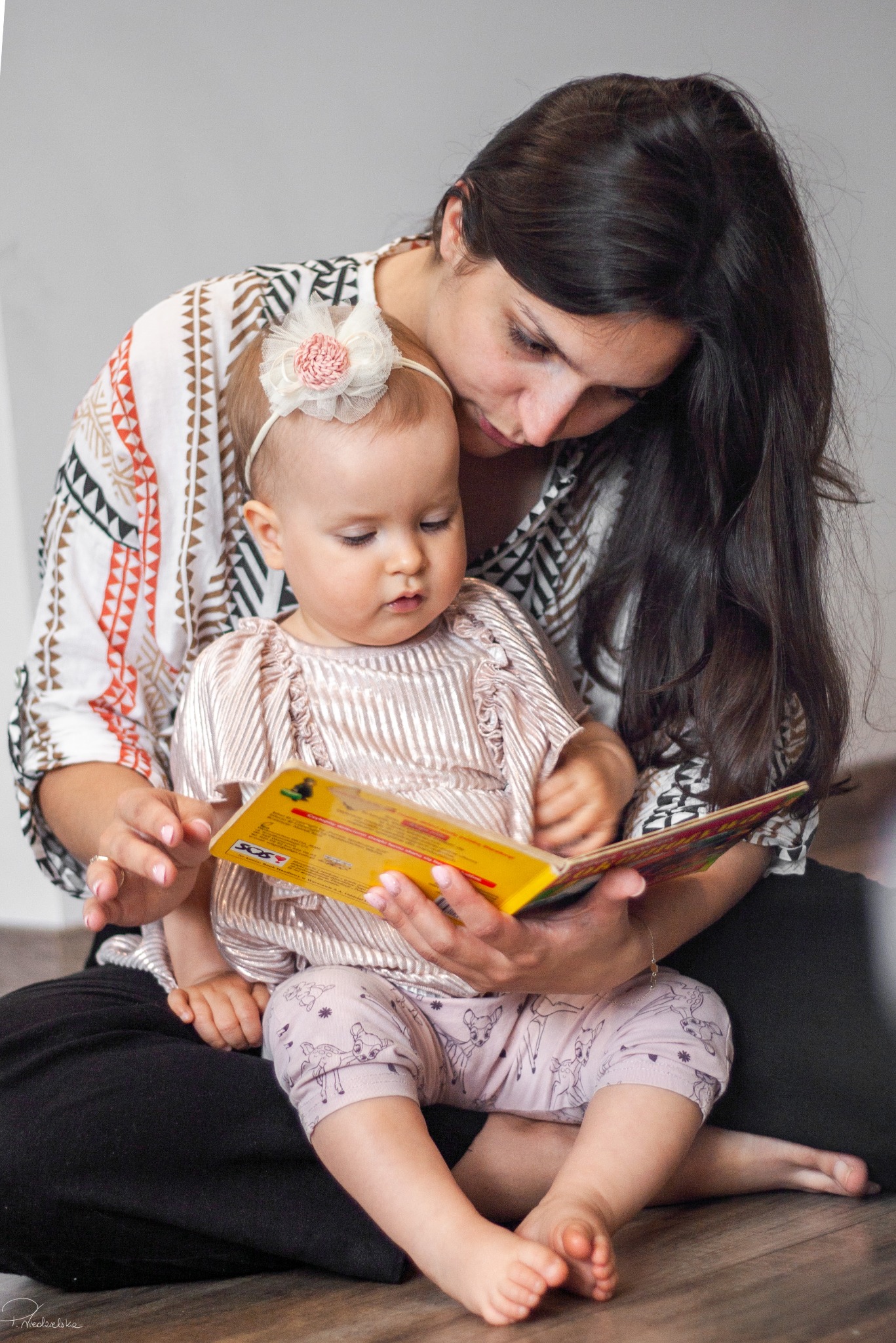 Matka z ciemnymi włosami czyta książkę córce z kokardą na głowie, siedząc na podłodze. Ubrana w bluzkę w etniczne wzory, dziecko w różowej bluzce i legginsach z motywem zwierzęcym.