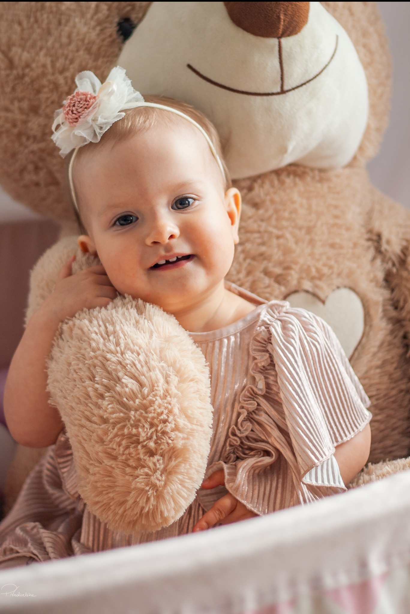 Urocza dziewczynka w pastelowej sukience z falbankami, przytulająca pluszowego misia z sercem, ozdobiona opaską z kwiatem. Delikatne światło, ciepłe kolory.