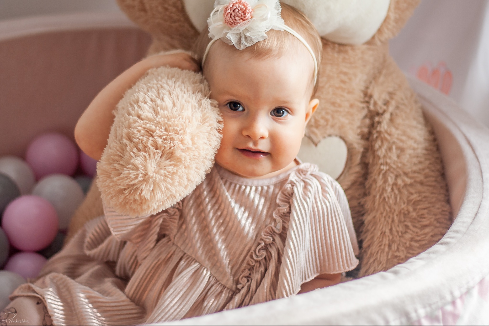 Urocza niemowlęca sesja: Dziewczynka w pudrowej sukience z pluszowym misiem w tle, otoczona kolorowymi piłeczkami, z delikatną opaską na głowie.