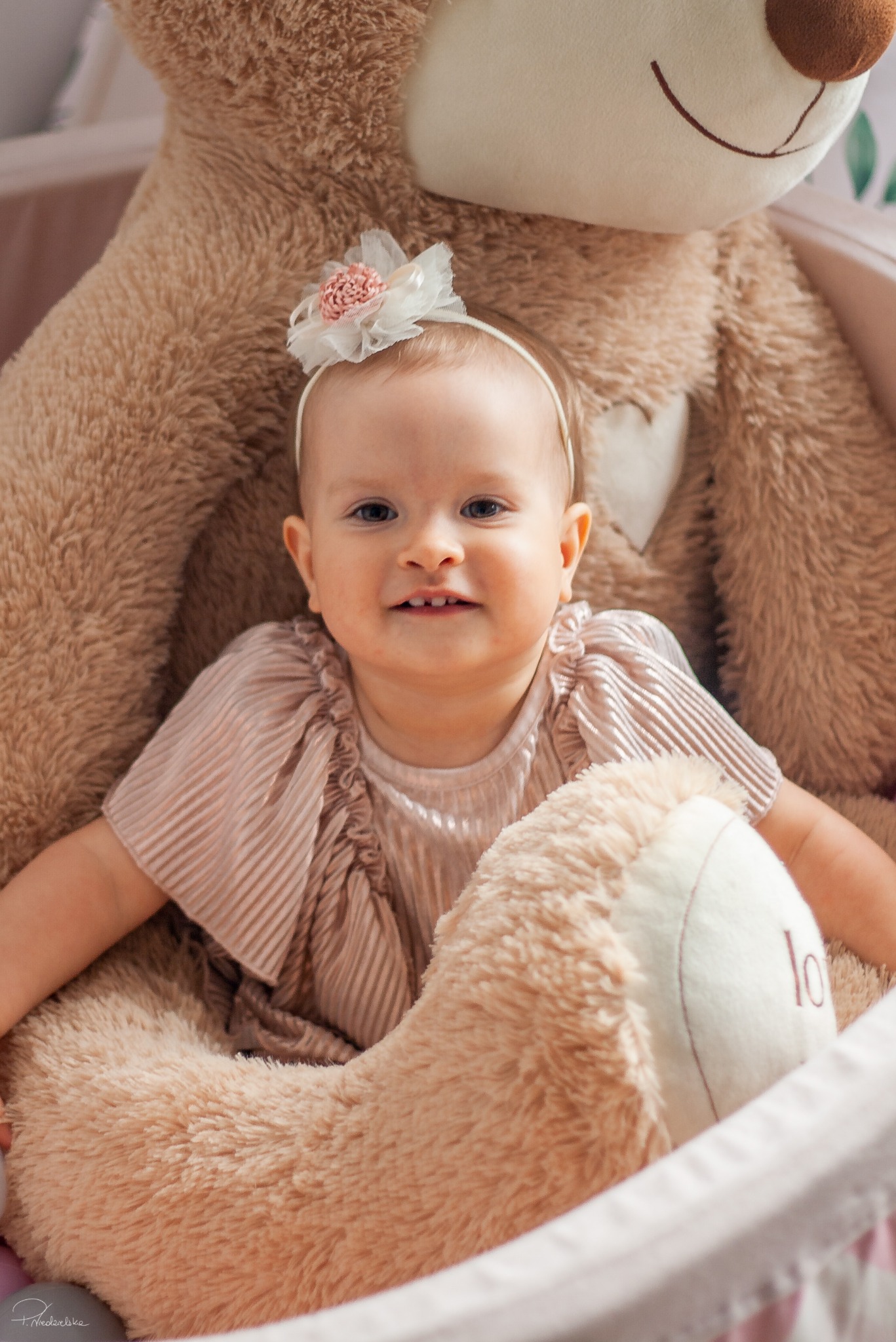 Uśmiechnięta dziewczynka z opaską na głowie, otoczona pluszowym misiem w łóżeczku, w ciepłych, naturalnych barwach. Portret dziecka w miękkim świetle.