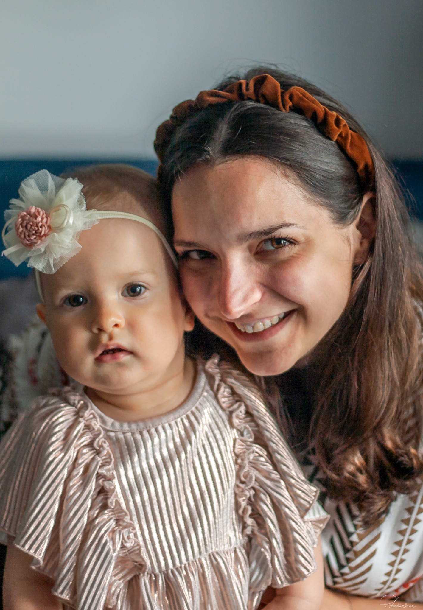 Portret mamy z córeczką, obie uśmiechnięte, w jasnym oświetleniu. Dziecko ma ozdobę we włosach w postaci kwiatka. Ujęcie od przodu, bliski plan.