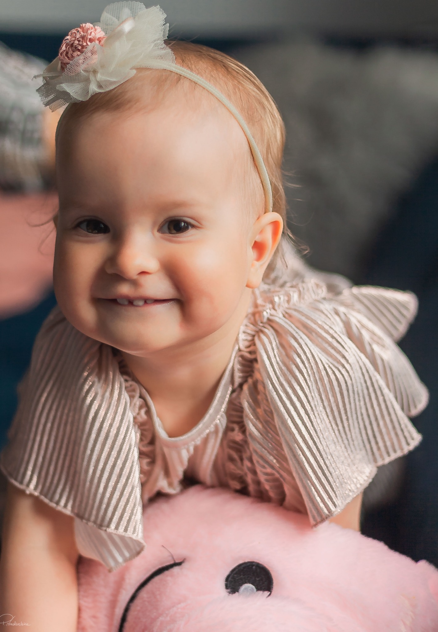 Uśmiechnięta dziewczynka w sukience w paski, z opaską z kwiatkiem, oparta na pluszowej śwince. Portret w ciepłych barwach, miękkie światło.