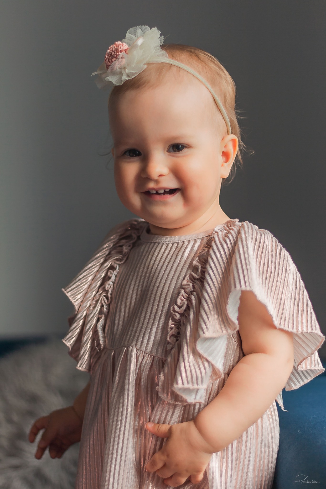 Uśmiechnięta dziewczynka w sukience z falbankami i opaską z kwiatkiem na głowie, na neutralnym tle. Delikatne światło podkreśla detale stroju.