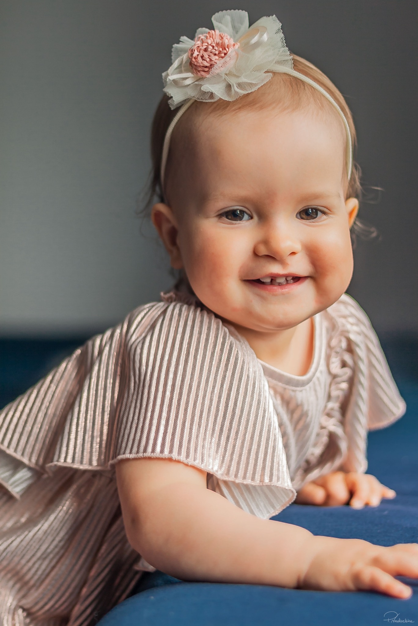 Uśmiechnięta dziewczynka w sukience w paski z kwiatową opaską na głowie, leżąca na niebieskim tle. Widoczny delikatny uśmiech i pierwsze ząbki.