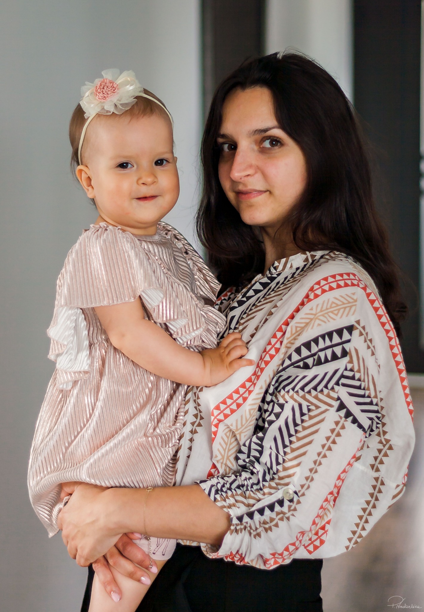 Portret matki z córeczką w eleganckich strojach. Dziewczynka ma ozdobę we włosach. Uśmiechnięte twarze, stonowana kolorystyka, naturalne światło.